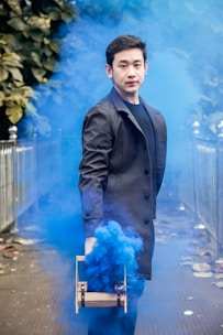A man in a black coat holds a device releasing blue smoke, surrounded by foliage and metal railings in an outdoor setting.