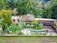 A lush tropical setting with a central thatched-roof building surrounded by dense greenery, including palm trees and bamboo. The area features a landscaped garden with a small curving pool. Nearby, there is a wooden deck and several open-air structures with draped curtains.