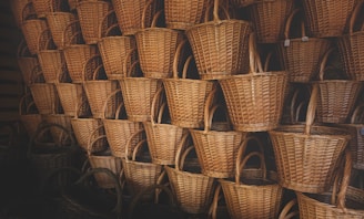A large collection of woven baskets stacked neatly in rows. The baskets are made of light brown wicker and have sturdy handles. They are arranged tightly together in a dimly lit space, creating a repetitive pattern.