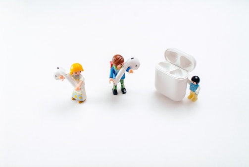 Three toy figures are positioned on a white background. Two of them are holding large earbud headphones, while the third stands next to an open case designed for storing the earbuds.
