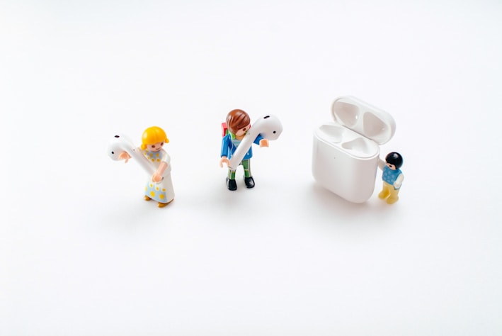 Three toy figures are positioned on a white background. Two of them are holding large earbud headphones, while the third stands next to an open case designed for storing the earbuds.
