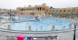 A large, ornate thermal bath complex with a bright yellow facade and arched windows surrounds a crowded outdoor pool. The pool is full of people swimming and relaxing, while some others are standing on the surrounding stone platforms. Benches with towels and personal items are visible in the foreground.
