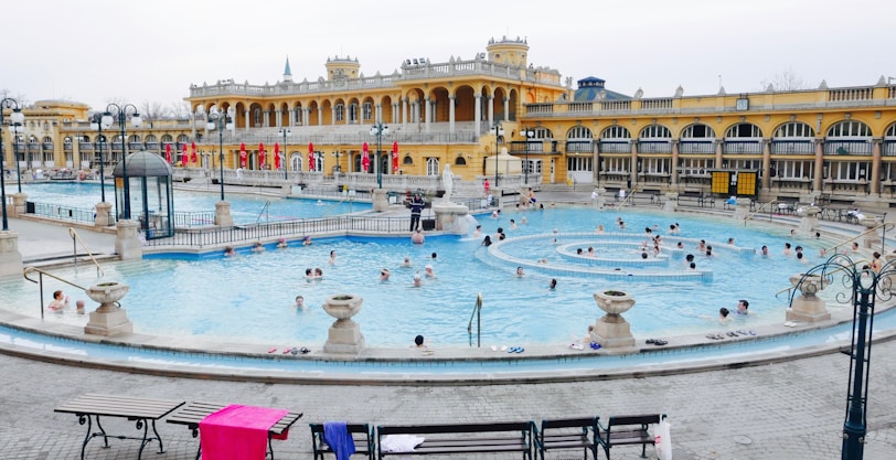 A large, ornate thermal bath complex with a bright yellow facade and arched windows surrounds a crowded outdoor pool. The pool is full of people swimming and relaxing, while some others are standing on the surrounding stone platforms. Benches with towels and personal items are visible in the foreground.