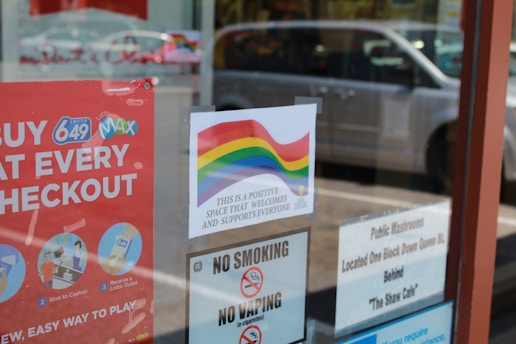 A storefront window features several signs. One sign displays a rainbow flag with the message 'This is a positive space that welcomes and supports everyone.' Nearby, there's a red sign promoting lottery games with instructions on how to play. Other signs include a 'No Smoking' and 'No Vaping' notice and a public restrooms location guide. In the reflection, a silver minivan and other vehicles can be seen parked outside.