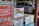 A storefront window features several signs. One sign displays a rainbow flag with the message 'This is a positive space that welcomes and supports everyone.' Nearby, there's a red sign promoting lottery games with instructions on how to play. Other signs include a 'No Smoking' and 'No Vaping' notice and a public restrooms location guide. In the reflection, a silver minivan and other vehicles can be seen parked outside.
