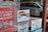 A storefront window features several signs. One sign displays a rainbow flag with the message 'This is a positive space that welcomes and supports everyone.' Nearby, there's a red sign promoting lottery games with instructions on how to play. Other signs include a 'No Smoking' and 'No Vaping' notice and a public restrooms location guide. In the reflection, a silver minivan and other vehicles can be seen parked outside.