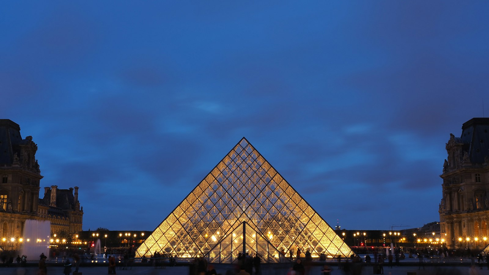 The Louvre's glass pyramid lit from within at blue hour, framed by the palace wings