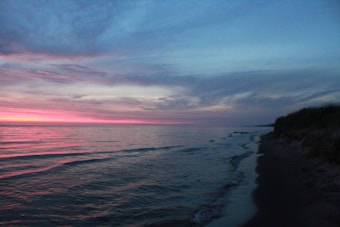 A tranquil beach scene features a gentle shoreline curving along the right with soft waves lapping at the sand. The sky is filled with a blend of blue and pink hues, suggesting a sunset or sunrise. The horizon is subtly highlighted with vibrant pink and purple tones, reflecting on the calm water surface.