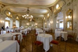 An elegant, ornate dining room with high ceilings, elaborate chandeliers, and intricately decorated walls. Round tables with white tablecloths are set for dining, and a few people are present, enhancing the ambience. The room features classical architectural elements and golden framed mirrors, adding to the luxurious feel.