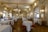 An elegant, ornate dining room with high ceilings, elaborate chandeliers, and intricately decorated walls. Round tables with white tablecloths are set for dining, and a few people are present, enhancing the ambience. The room features classical architectural elements and golden framed mirrors, adding to the luxurious feel.