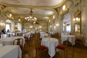 An elegant, ornate dining room with high ceilings, elaborate chandeliers, and intricately decorated walls. Round tables with white tablecloths are set for dining, and a few people are present, enhancing the ambience. The room features classical architectural elements and golden framed mirrors, adding to the luxurious feel.
