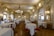 An elegant, ornate dining room with high ceilings, elaborate chandeliers, and intricately decorated walls. Round tables with white tablecloths are set for dining, and a few people are present, enhancing the ambience. The room features classical architectural elements and golden framed mirrors, adding to the luxurious feel.