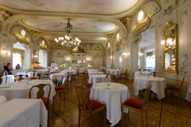 An elegant, ornate dining room with high ceilings, elaborate chandeliers, and intricately decorated walls. Round tables with white tablecloths are set for dining, and a few people are present, enhancing the ambience. The room features classical architectural elements and golden framed mirrors, adding to the luxurious feel.