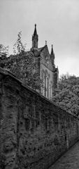 A gothic-style building with ornate spires and arched windows stands partially obscured by trees. A stone wall leads the eye towards the building, creating a sense of direction and depth. The overcast sky adds a moody atmosphere to the scene.