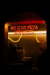 A dimly lit interior with a neon sign displaying the words 'WE LOVE PIZZA SPAGHETTI' in bright red and yellow lights. There are tables and chairs visible, suggesting a cozy, intimate dining space.