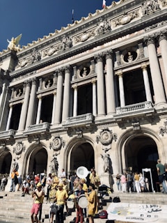 A group of musicians dressed in bright yellow clothes perform on the steps of an ornate historic building. The architecture features tall columns, intricate carvings, and statues. People are gathered around, enjoying the performance under a clear blue sky.