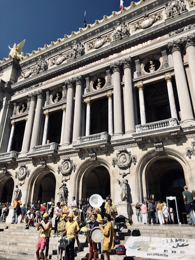 A group of musicians dressed in bright yellow clothes perform on the steps of an ornate historic building. The architecture features tall columns, intricate carvings, and statues. People are gathered around, enjoying the performance under a clear blue sky.