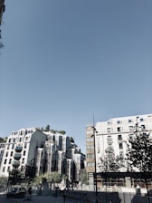 A modern urban setting featuring unique architectural buildings with reflective glass surfaces and greenery on rooftops. The sky is clear and blue, and there are trees and bicycles at ground level. A sign with 'NOVOTEL' is visible on one building.