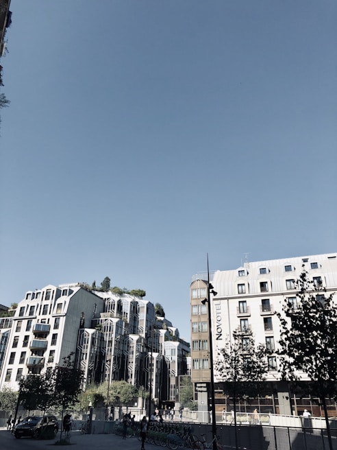 A modern urban setting featuring unique architectural buildings with reflective glass surfaces and greenery on rooftops. The sky is clear and blue, and there are trees and bicycles at ground level. A sign with 'NOVOTEL' is visible on one building.