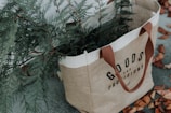 A jute tote bag with brown handles labeled 'Goods and Provisions', containing green fir branches. The bag is placed on a surface scattered with dry, brown leaves.