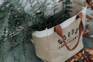 A jute tote bag with brown handles labeled 'Goods and Provisions', containing green fir branches. The bag is placed on a surface scattered with dry, brown leaves.
