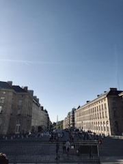 A bustling city street with classic European architecture, showing rows of historic buildings under a clear blue sky. In the distance, the Eiffel Tower is visible, while numerous people walk along the cobblestone streets beneath the sunlight.