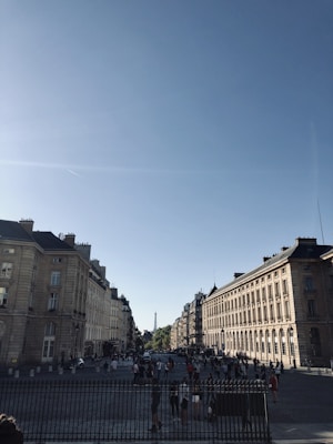 A bustling city street with classic European architecture, showing rows of historic buildings under a clear blue sky. In the distance, the Eiffel Tower is visible, while numerous people walk along the cobblestone streets beneath the sunlight.