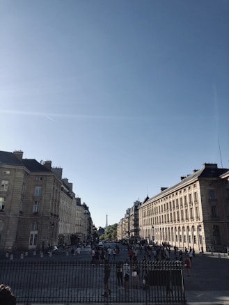 A bustling city street with classic European architecture, showing rows of historic buildings under a clear blue sky. In the distance, the Eiffel Tower is visible, while numerous people walk along the cobblestone streets beneath the sunlight.