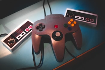 A retro gaming setup with a central gaming controller featuring multiple buttons and joysticks, flanked by two classic-style controllers with a simple directional pad and buttons. The background is a soft, blue surface giving a nostalgic feel.