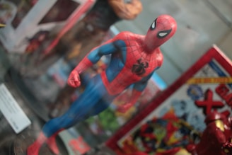 A detailed figurine of a superhero in a red and blue costume with a black spider emblem on the chest. The figure stands in a dynamic pose with one arm slightly forward. Surrounding it are various comic-themed items, including a book with vibrant illustrations.