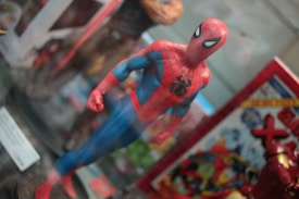 A detailed figurine of a superhero in a red and blue costume with a black spider emblem on the chest. The figure stands in a dynamic pose with one arm slightly forward. Surrounding it are various comic-themed items, including a book with vibrant illustrations.
