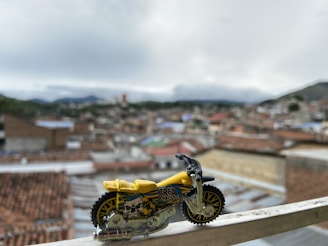 A small yellow and black toy motorcycle with intricate design details. It is placed on a metal railing with a blurred urban landscape in the background, consisting of rooftops and distant mountains under a cloudy sky.