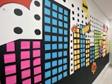 A vibrant wall display features a cityscape made of colorful rectangular shapes representing buildings, each with windows created from colored paper. Comic book-style elements, including speech bubbles with words like 'POW' and 'BOOM,' add a dynamic feel. Large black dots and a yellow symbol enhance the comic theme.