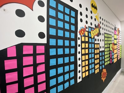 A vibrant wall display features a cityscape made of colorful rectangular shapes representing buildings, each with windows created from colored paper. Comic book-style elements, including speech bubbles with words like 'POW' and 'BOOM,' add a dynamic feel. Large black dots and a yellow symbol enhance the comic theme.