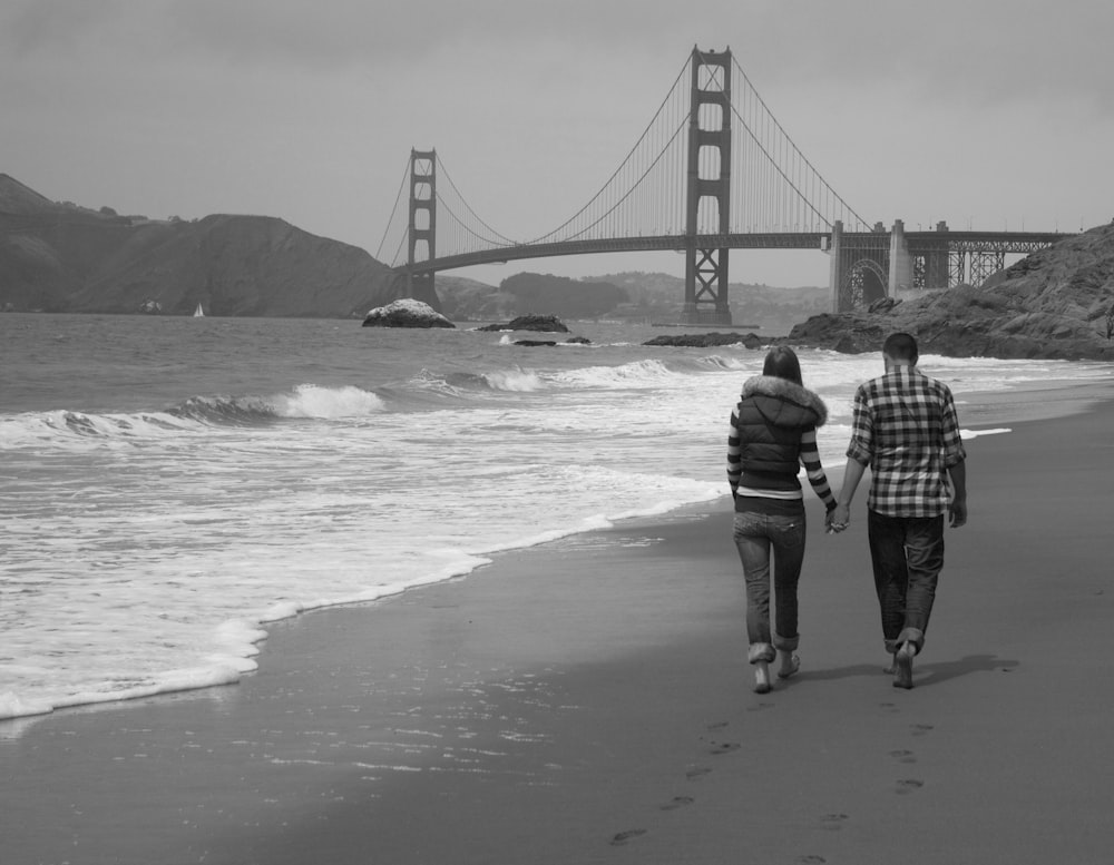 grayscale photography of man and woman walking near seashore viewing Golden Gate bridge
