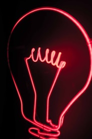 A glowing red neon light shaped like a classic light bulb against a dark background. The design is simple yet striking, with the filament represented by a zigzag pattern.