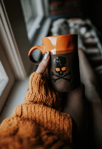 Close-up of a hand holding a bright orange stoneware cup with a cozy knitted blanket in the background.