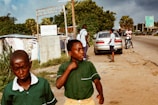 Two boys in green school uniforms are walking along a roadside. Behind them, a few people are standing near a parked car. A motorcyclist is also visible on the road. The area is lined with trees and some makeshift structures.