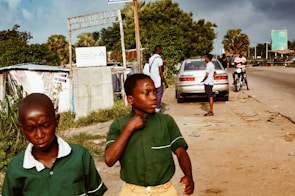 Two boys in green school uniforms are walking along a roadside. Behind them, a few people are standing near a parked car. A motorcyclist is also visible on the road. The area is lined with trees and some makeshift structures.