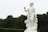 A classical white marble statue of a male figure holding a cup in one hand, with a draped cloth around his waist. The statue is adorned with detailed carvings of flowers and fruit and is set against a backdrop of green trees and cloudy sky.