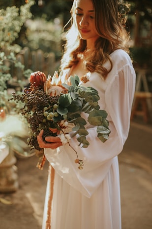 A soft-lit photo of a woman in a flowing peach dress holding a bouquet of wildflowers in a sun-dappled garden.