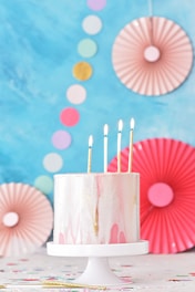 A pastel-colored cake with a marbled pink and white frosting, topped with four lit candles, is placed on a white cake stand. Decorative circular paper fans and multicolored dots adorn the background, which is primarily blue, creating a festive atmosphere.