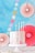 A pastel-colored cake with a marbled pink and white frosting, topped with four lit candles, is placed on a white cake stand. Decorative circular paper fans and multicolored dots adorn the background, which is primarily blue, creating a festive atmosphere.