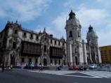 A large ornate colonial-style building with intricate stonework and wooden balconies is situated on a bustling urban street. People are walking across the crosswalk, and a few cars are visible on the road. The sky is partly cloudy, casting soft light on the scene.
