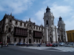A large ornate colonial-style building with intricate stonework and wooden balconies is situated on a bustling urban street. People are walking across the crosswalk, and a few cars are visible on the road. The sky is partly cloudy, casting soft light on the scene.
