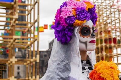 A person with their face painted in the traditional style of Dia de los Muertos, with intricate designs and dark eye sockets. They are wearing a vibrant floral headdress made up of purple, pink, and orange flowers, and holding a bouquet of similar colored flowers. In the background, colorful decorations and festive structures add to the celebratory atmosphere.