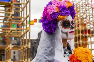 A person with their face painted in the traditional style of Dia de los Muertos, with intricate designs and dark eye sockets. They are wearing a vibrant floral headdress made up of purple, pink, and orange flowers, and holding a bouquet of similar colored flowers. In the background, colorful decorations and festive structures add to the celebratory atmosphere.