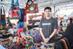 A person stands smiling behind a colorful market stall displaying a variety of handcrafted items. The stall is filled with vibrant bags, textiles, and small handcrafted objects, showcasing an array of traditional designs and patterns. The setting is a busy, indoor market area with other vendors and visitors in the background.