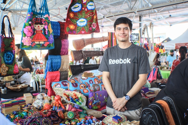 A person stands smiling behind a colorful market stall displaying a variety of handcrafted items. The stall is filled with vibrant bags, textiles, and small handcrafted objects, showcasing an array of traditional designs and patterns. The setting is a busy, indoor market area with other vendors and visitors in the background.