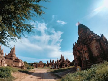 A series of ancient temples and pagodas made of brown brick are situated under a bright blue sky with scattered white clouds. The scene is set on a dusty path surrounded by green vegetation and large trees. The sunlight creates a serene and timeless atmosphere.